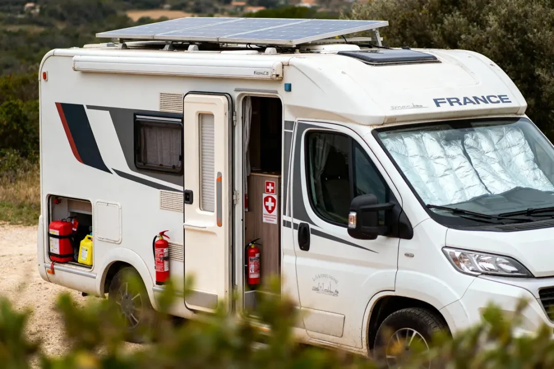 Camping-car blanc avec panneaux solaires, extincteurs, et trousse de secours. Stationné dans un paysage naturel.