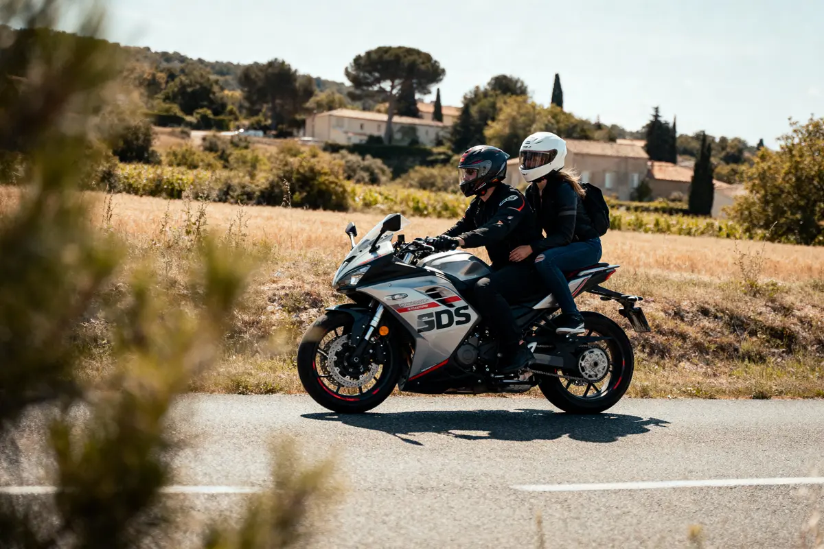 Deux personnes en moto grise sur route de campagne ensoleillée, arbres et maison en arrière-plan.
