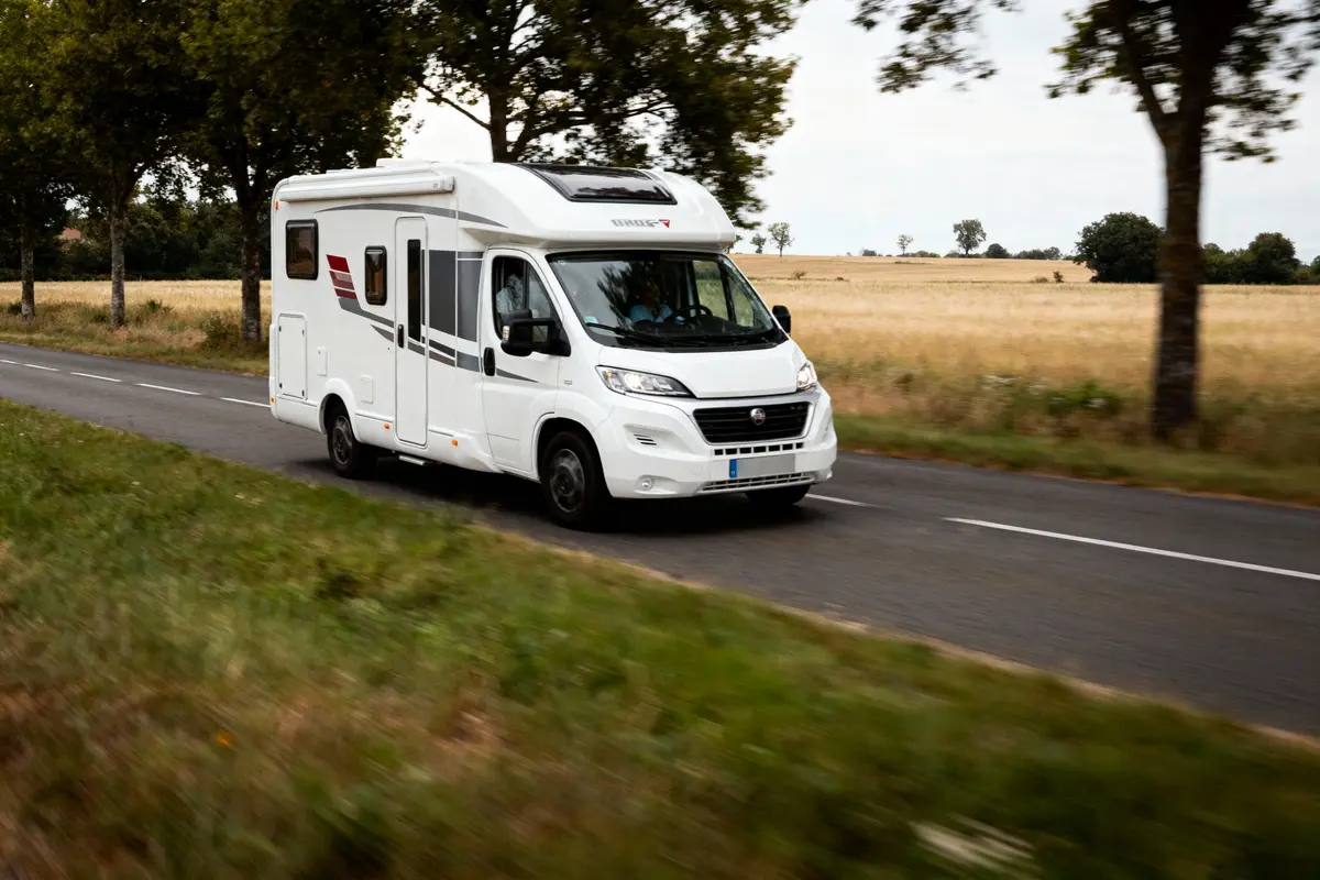 Camping-car blanc roulant sur une route bordée d'arbres et de champs verdoyants, sous un ciel partiellement nuageux.