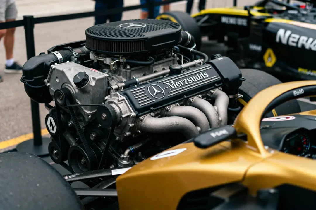 Gros plan d'un moteur automobile haute performance écrit "Merzsudlis", avec voitures de course en arrière-plan.