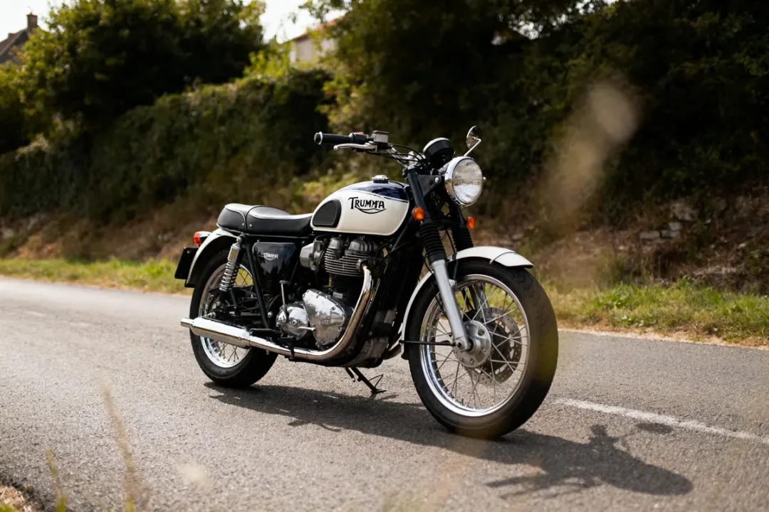 Moto vintage Triumph garée sur une route, entourée de verdure. Modèle classique avec éléments chromés.