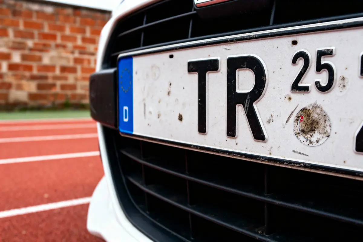 Plaque d'immatriculation européenne sale sur une voiture blanche, route en briques et asphalte rouge en arrière-plan.
