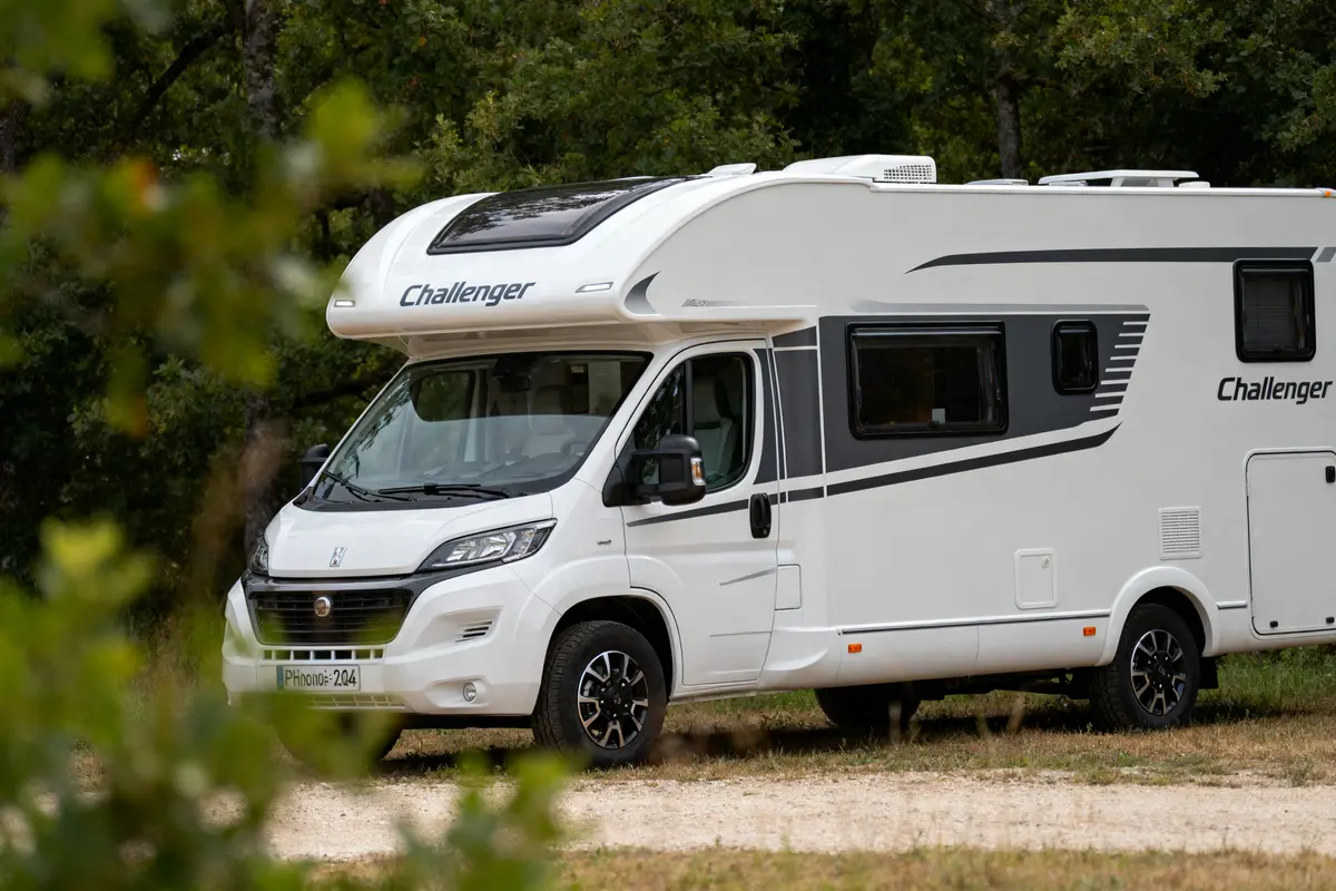 Camping-car blanc de marque Challenger garé dans un cadre verdoyant, partiellement caché par des branches.