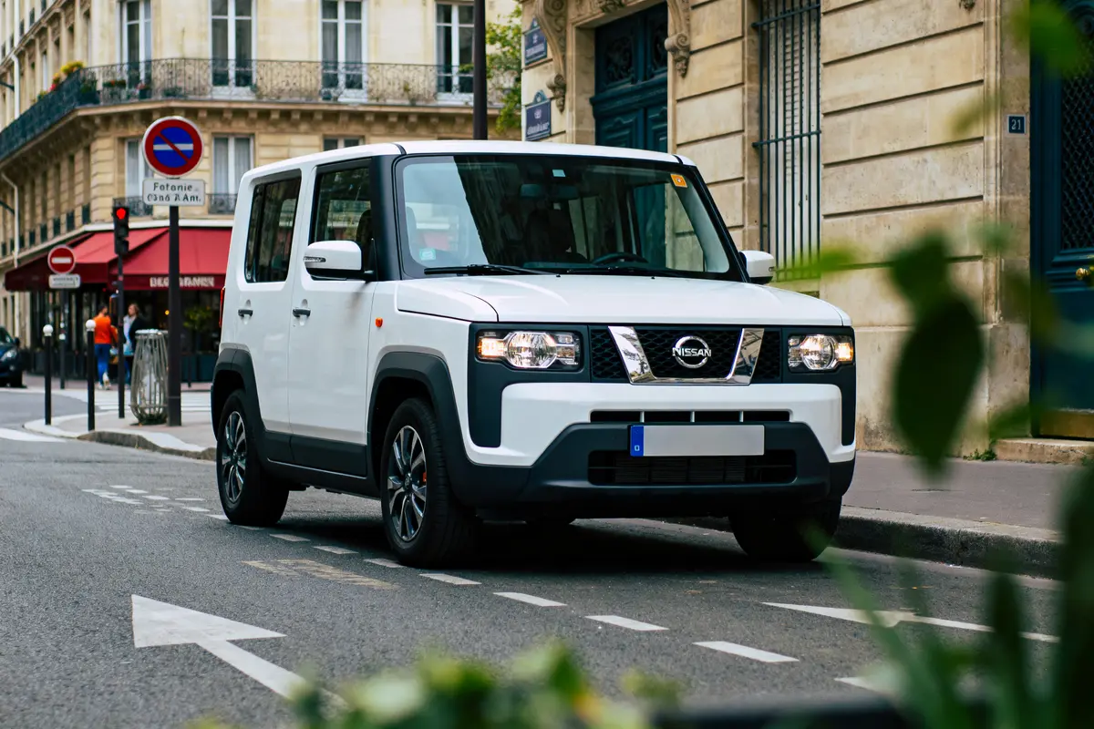 Voiture Nissan blanche sur rue urbaine, immeubles historiques en arrière-plan, végétation floue au premier plan.