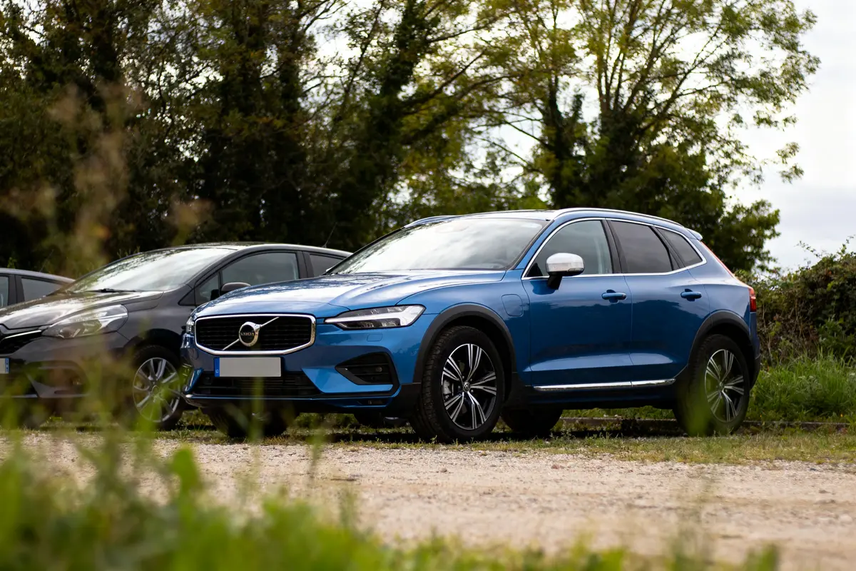 Voiture bleue Volvo stationnée à l'extérieur, à côté d'un véhicule noir, arbres en arrière-plan.