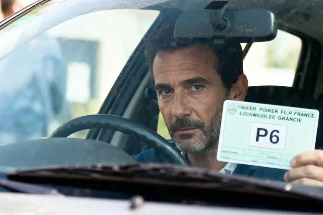 Homme dans une voiture montrant un document avec "P6" à travers la fenêtre avant, expression sérieuse.