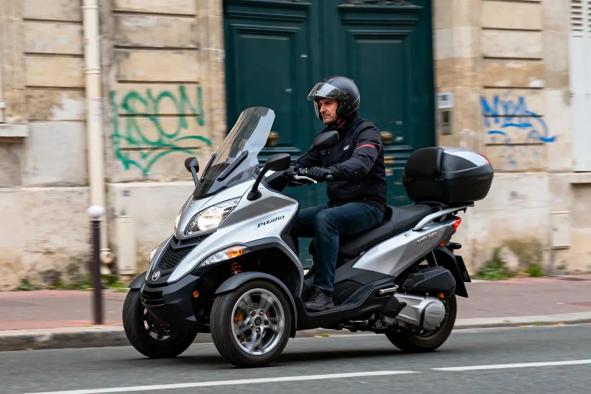 Homme conduisant un scooter Piaggio trois-roues argenté en ville, portant un casque noir.