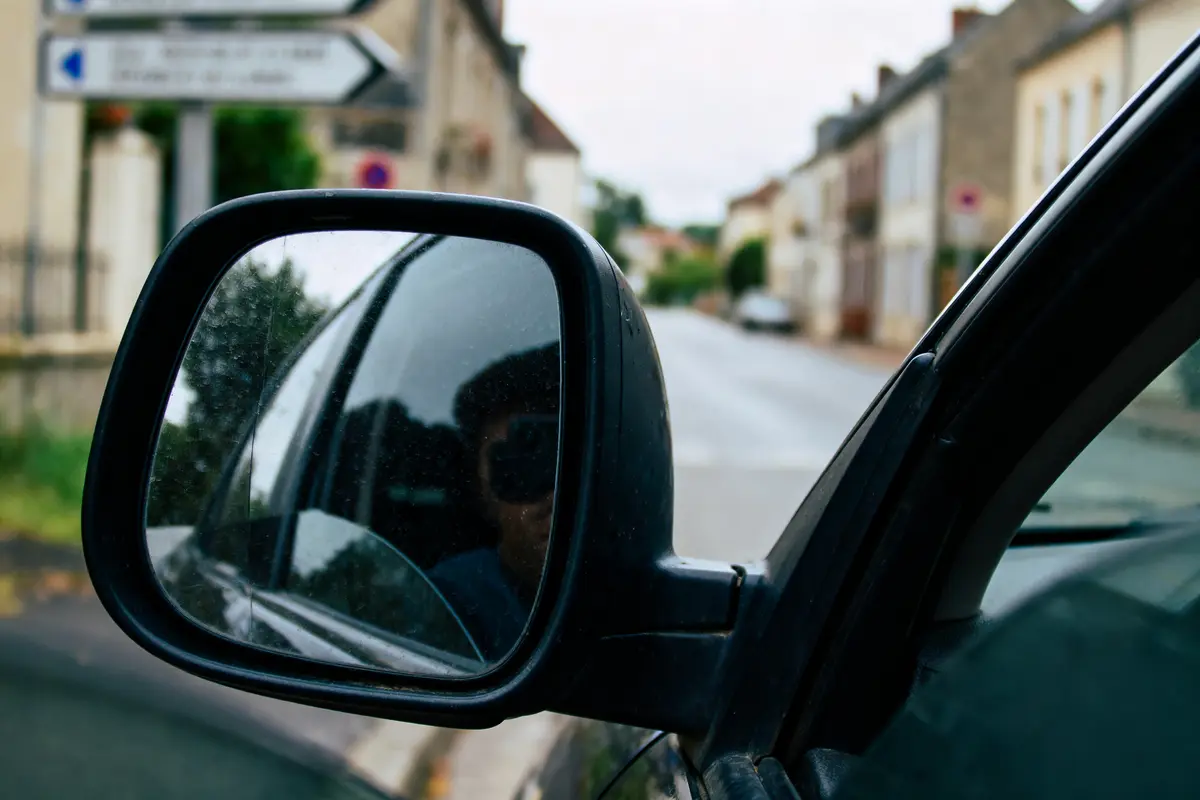 Rétroviseur de voiture reflétant rue, maisons floues en arrière-plan, panneau signalisation visible côté gauche.