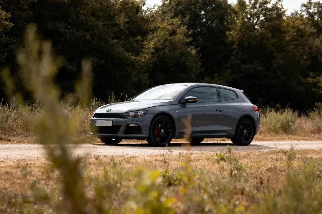 Voiture grise sur chemin de terre, entourée de végétation, avec forêt en arrière-plan par temps ensoleillé.