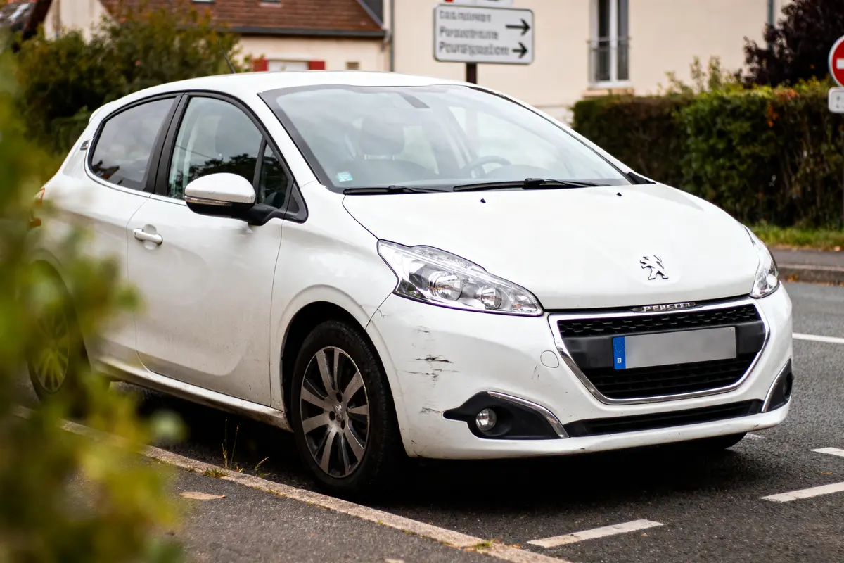 Voiture blanche Peugeot garée, légèrement éraflée à l'avant, avec panneau directionnel en arrière-plan.