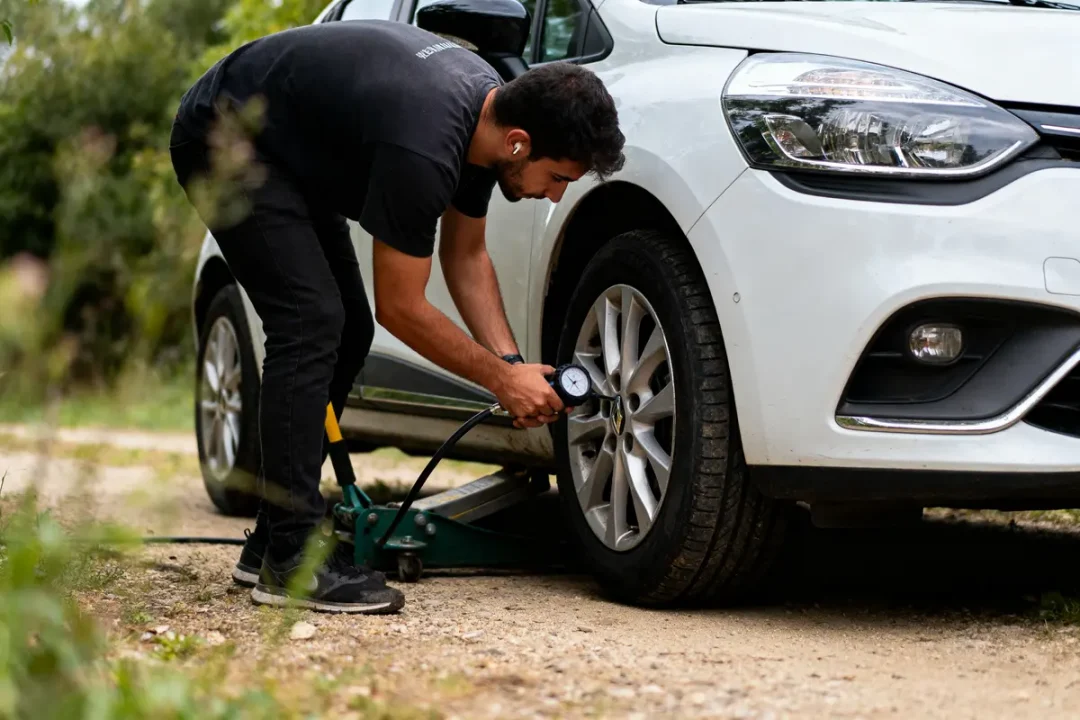 Homme vérifiant la pression d'un pneu de voiture blanche avec manomètre portable, extérieur, nature autour.