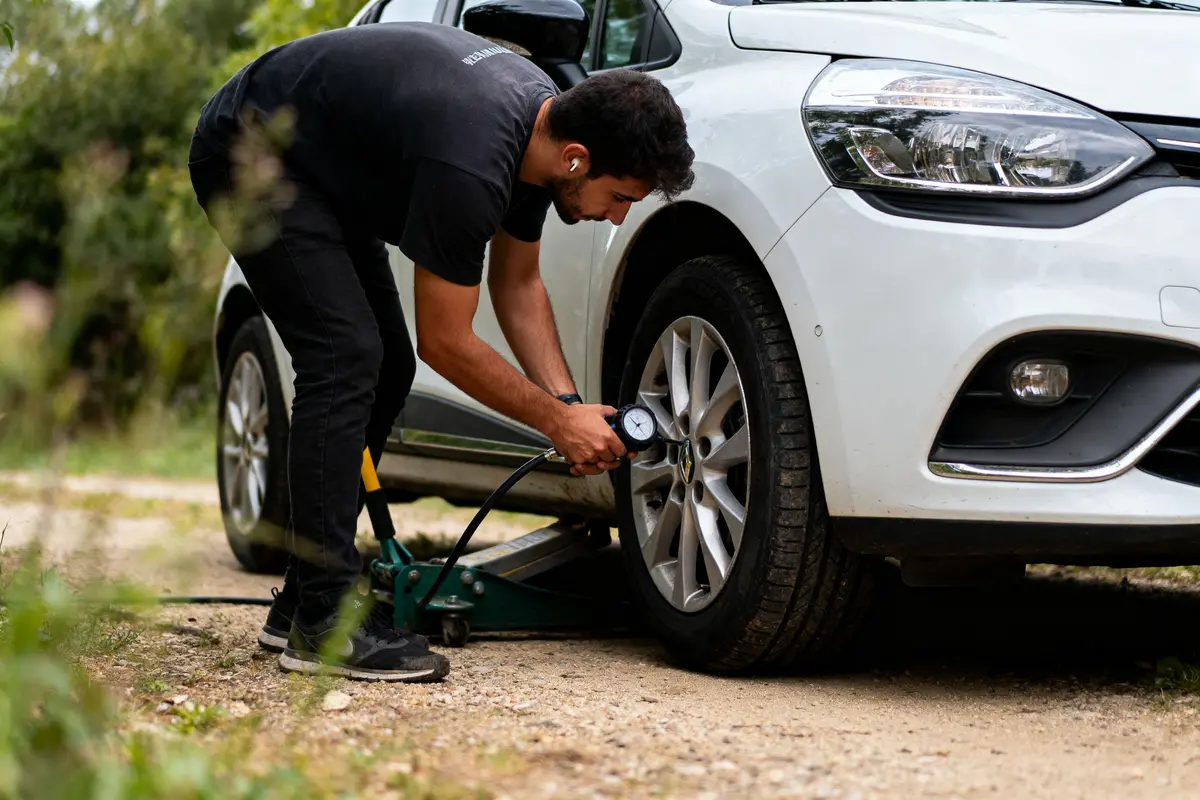 Homme vérifiant la pression d'un pneu de voiture blanche avec manomètre portable, extérieur, nature autour.
