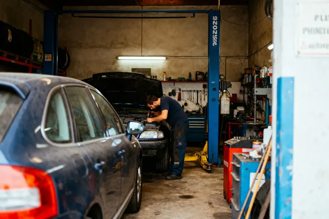 Mécanicien réparant une voiture dans un garage, outils suspendus au mur, véhicule aux phares ouverts.