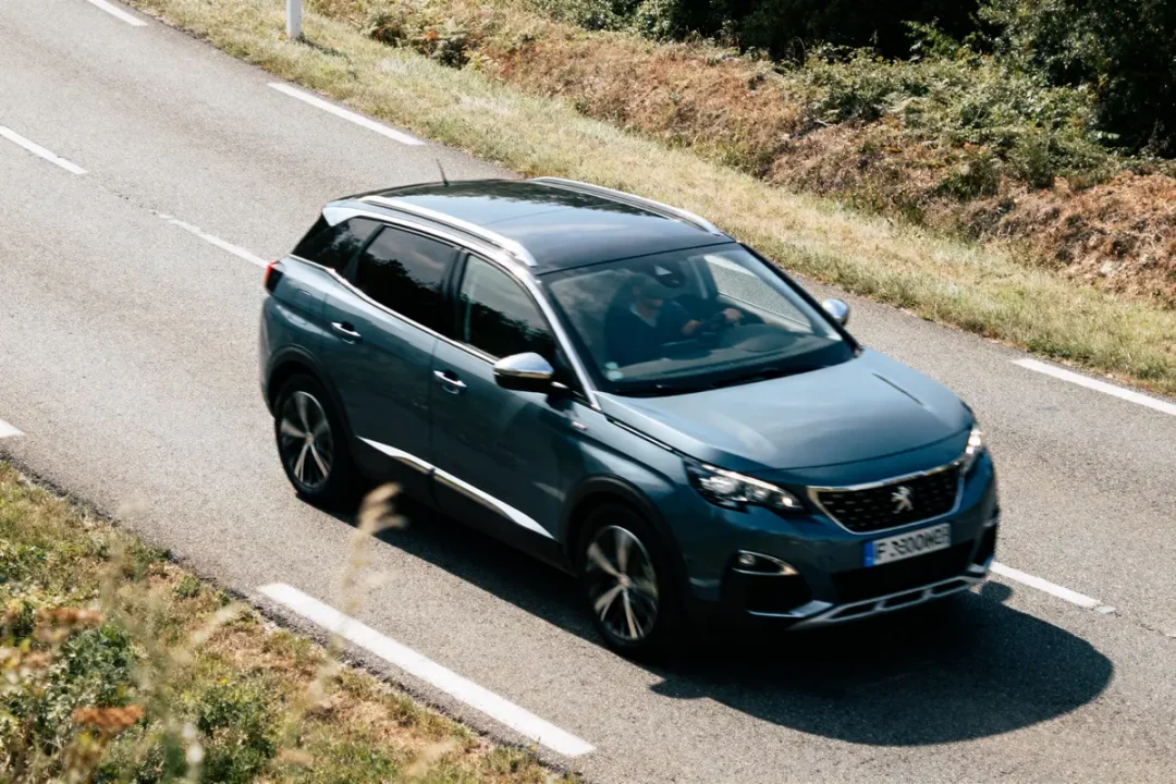 SUV bleu roulant sur une route de campagne bordée d'herbe et d'arbustes.