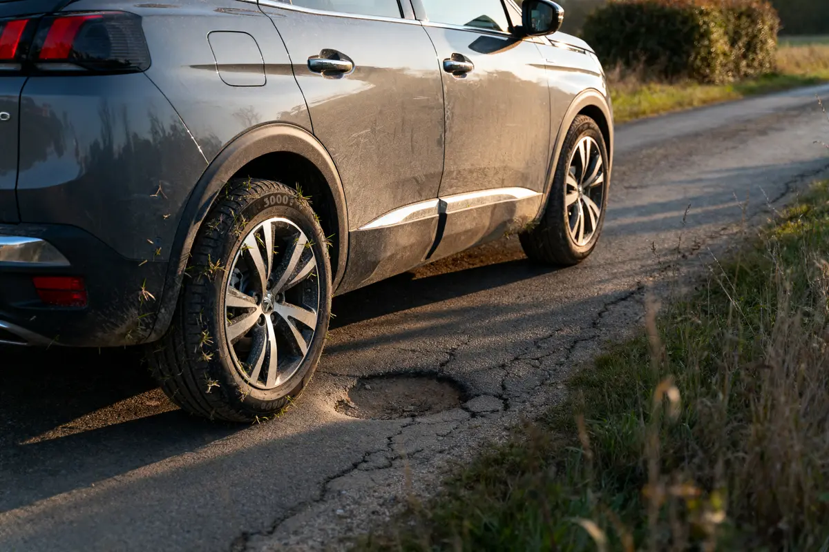 SUV gris sur route endommagée avec des nids-de-poule et herbes sur le bas-côté. Pneus sales.