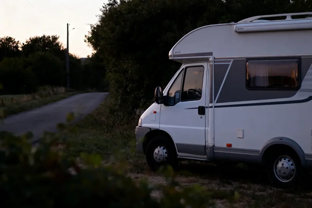 Camping-car blanc garé au bord d'une route de campagne, au crépuscule, entouré de végétation.