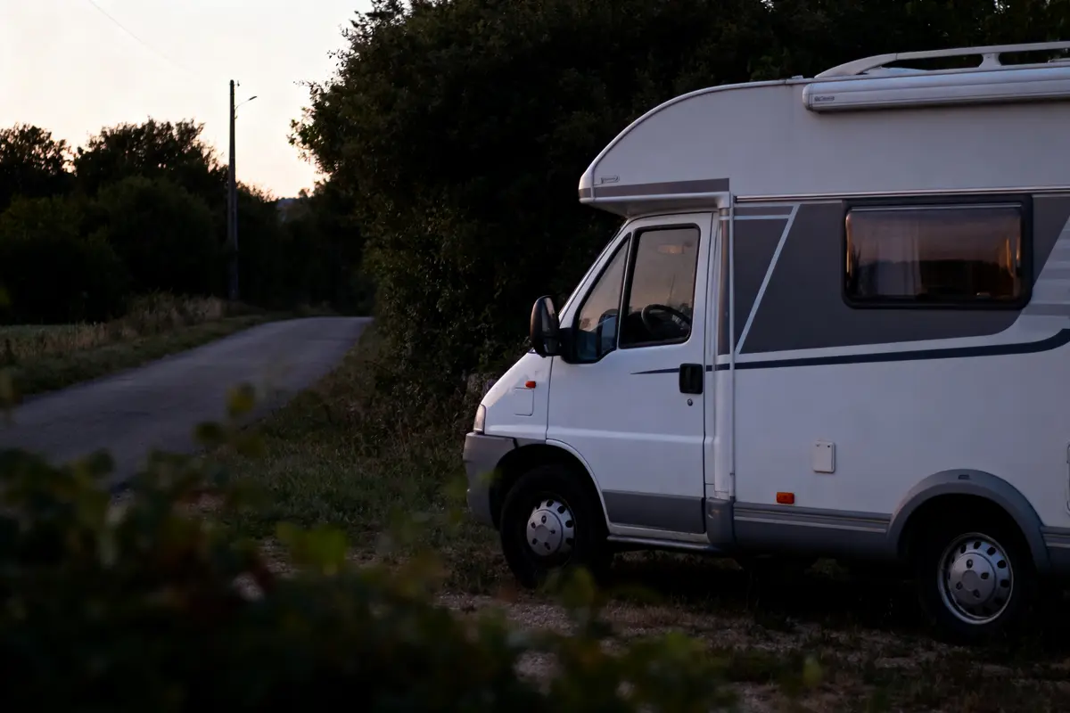 Camping-car blanc garé au bord d'une route de campagne, au crépuscule, entouré de végétation.