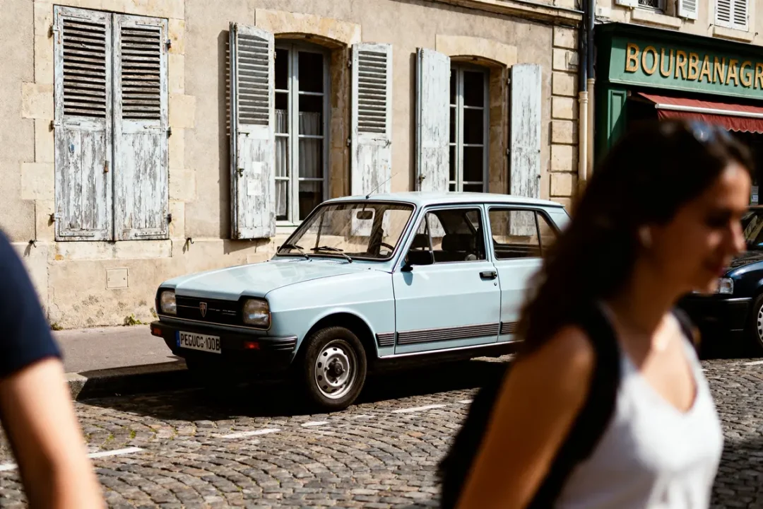 Voiture classique bleue garée devant bâtiment ancien, fenêtres à volets. Personne floue passe devant.