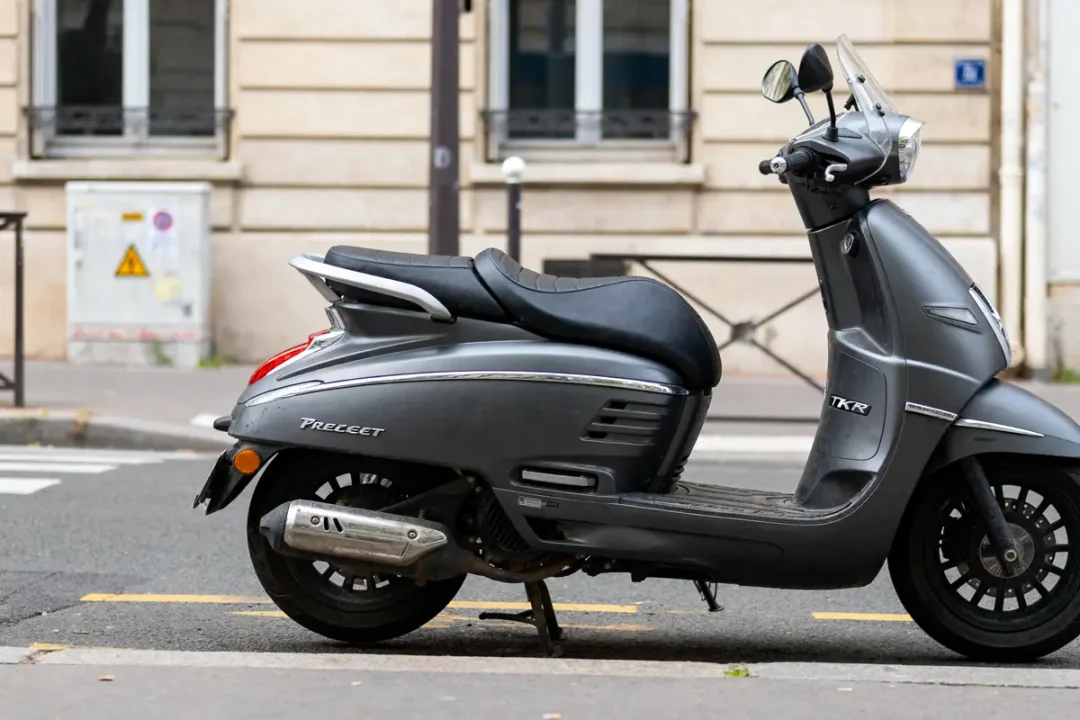 Scooter gris stationné dans la rue devant un bâtiment en pierre avec fenêtres et boîte électrique.