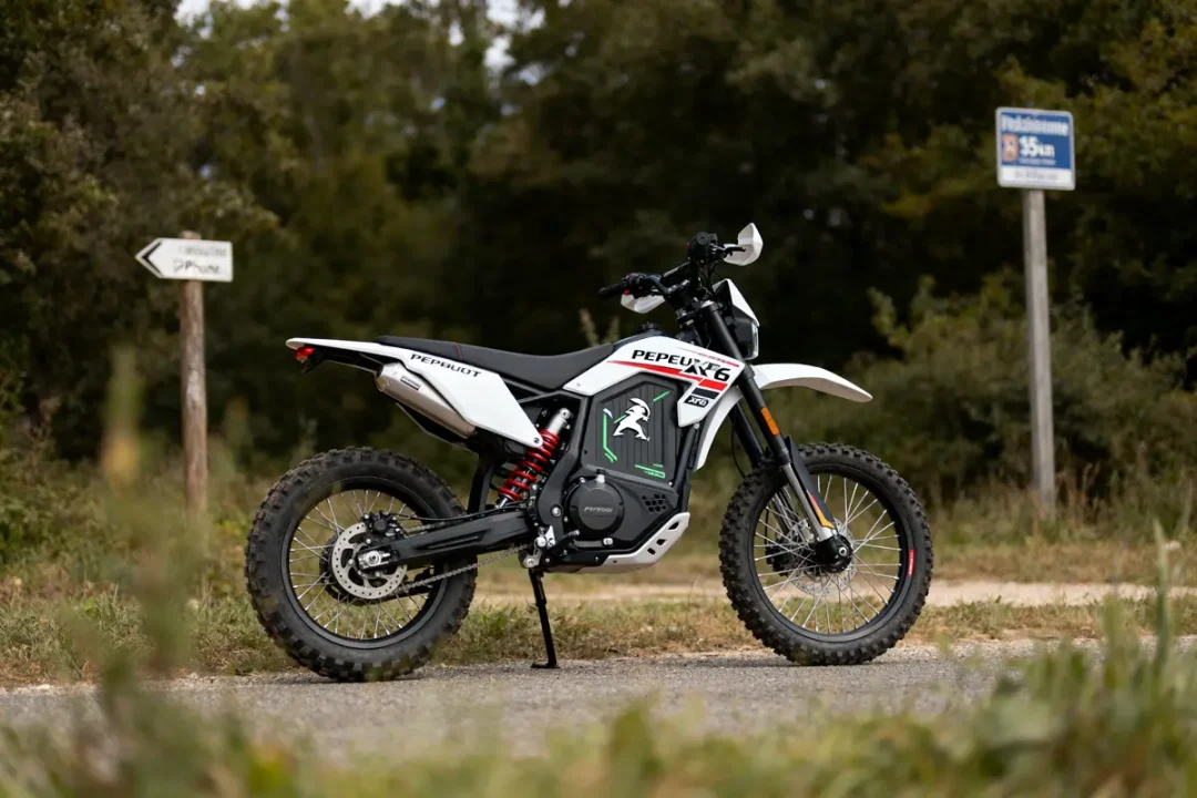 Moto tout-terrain blanche sur chemin forestier, panneaux de signalisation en arrière-plan, journée ensoleillée.