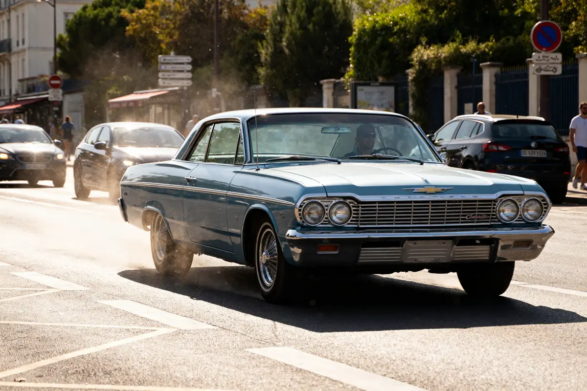 Un Chevrolet Impala bleu roule sur une route bordée de voitures et d'arbres
